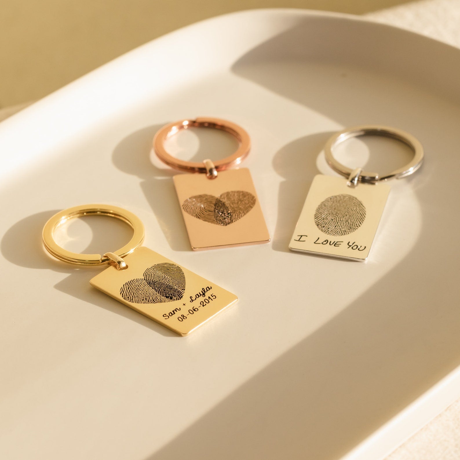 Three rectangular keychains with fingerprints engraved on them, displayed in a triangular arrangement on a light surface. The keychains have options of different finishes, including gold and rose gold.